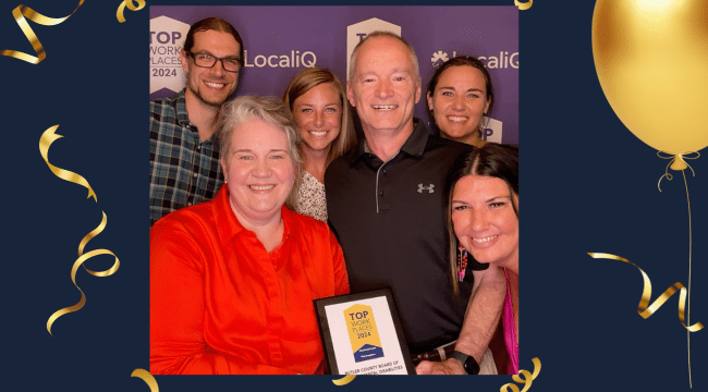 A group of people squeeze together for a picture. The woman in front holds a plaque that shows the Top Workplaces logo and some indistinguishable text.